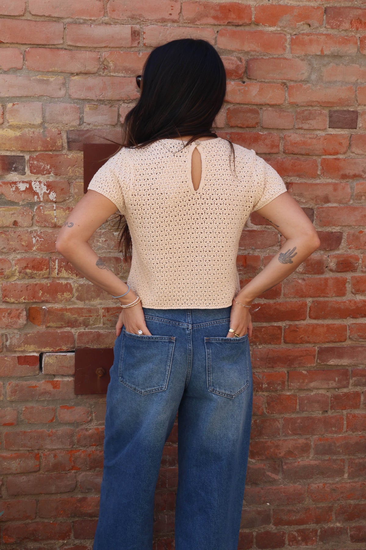 Person wearing a beige crochet top and blue jeans standing against a brick wall.