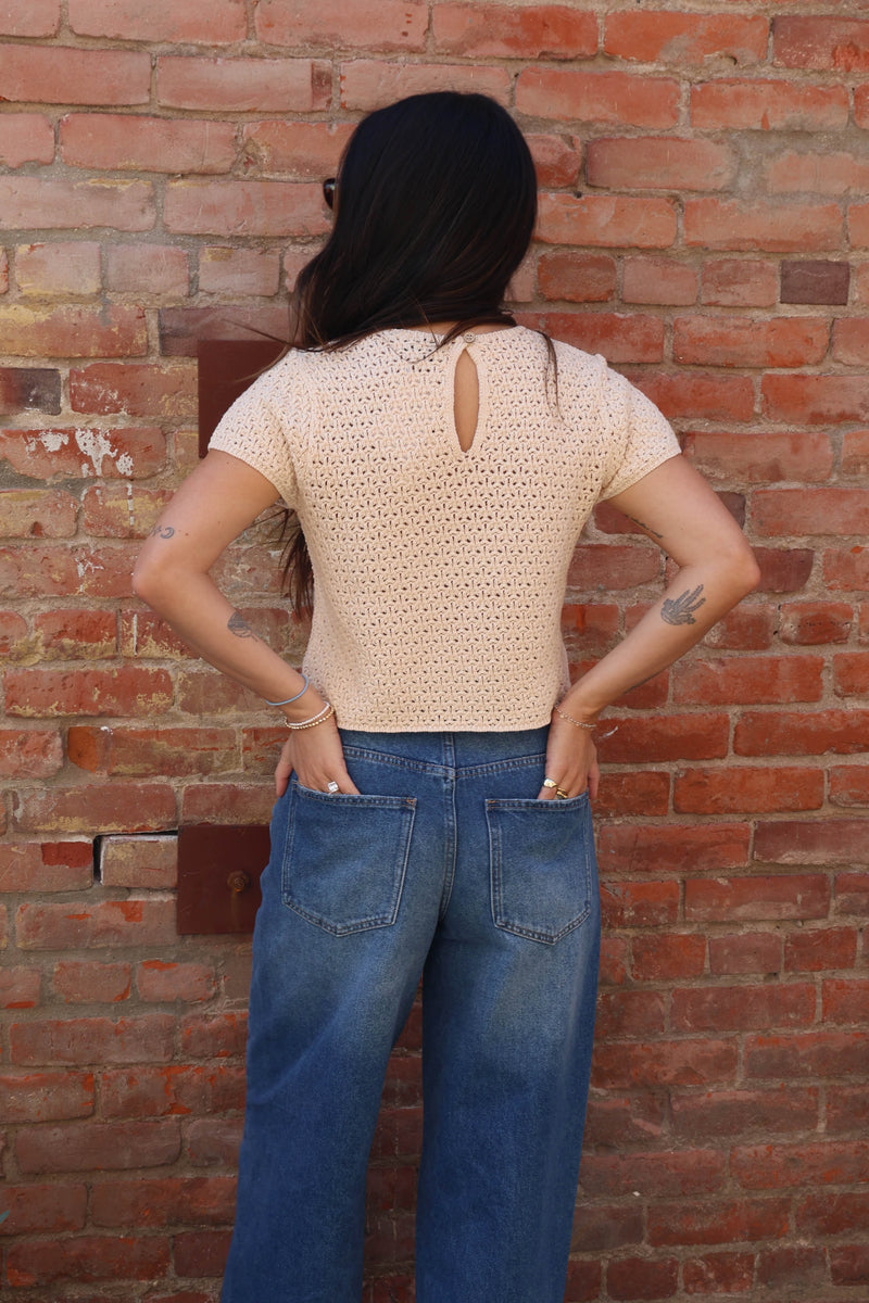Person wearing a beige crochet top and blue jeans standing against a brick wall.