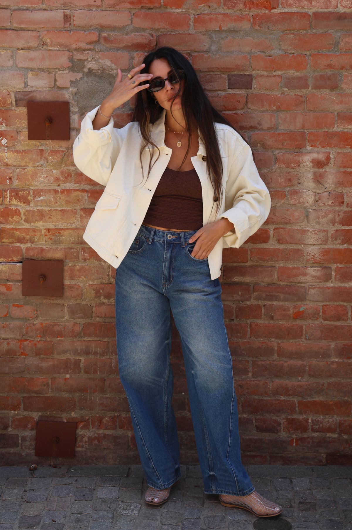 Woman in a white jacket and blue jeans standing against a brick wall.