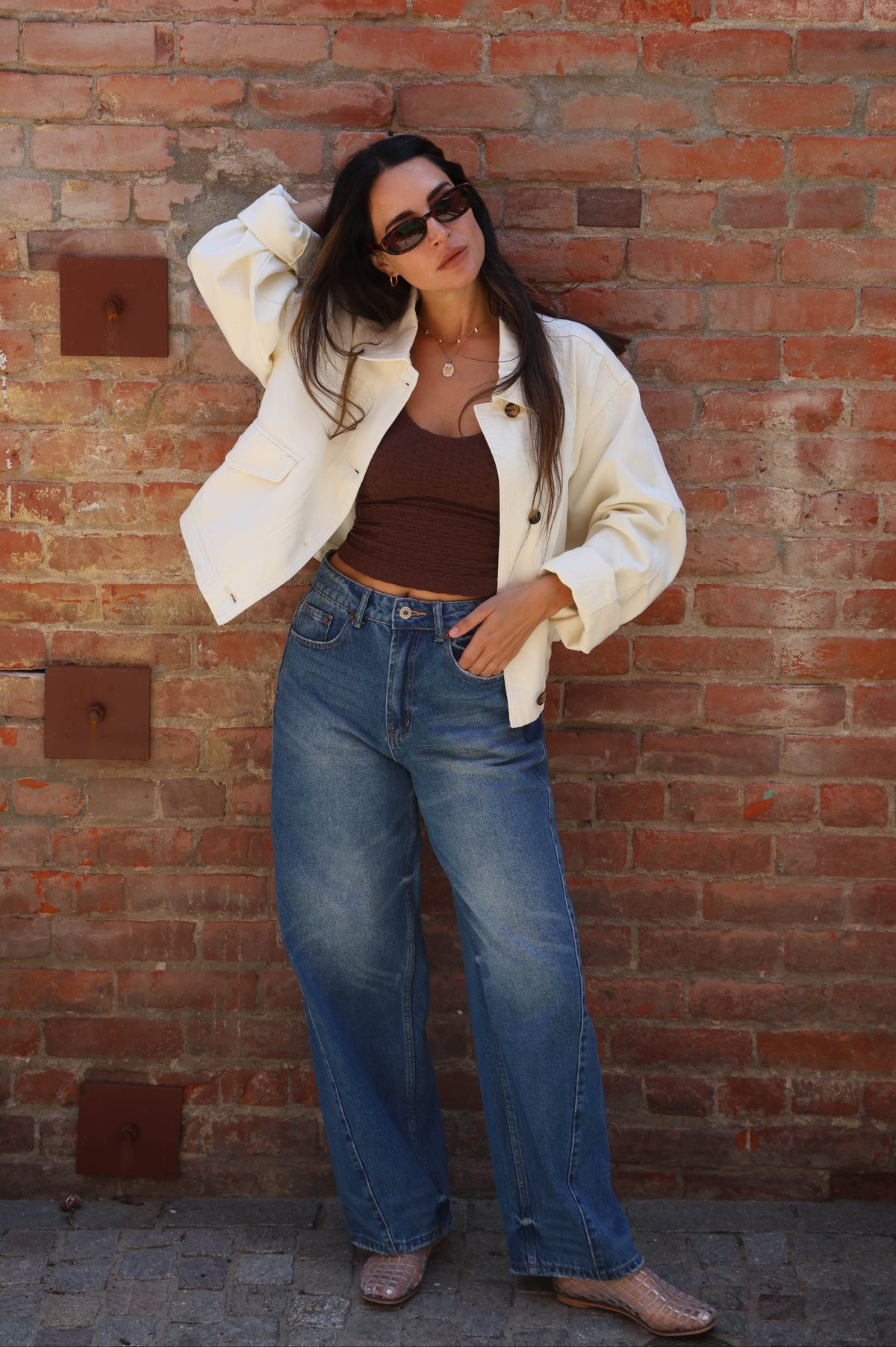 Woman in a white jacket and blue jeans standing against a brick wall.