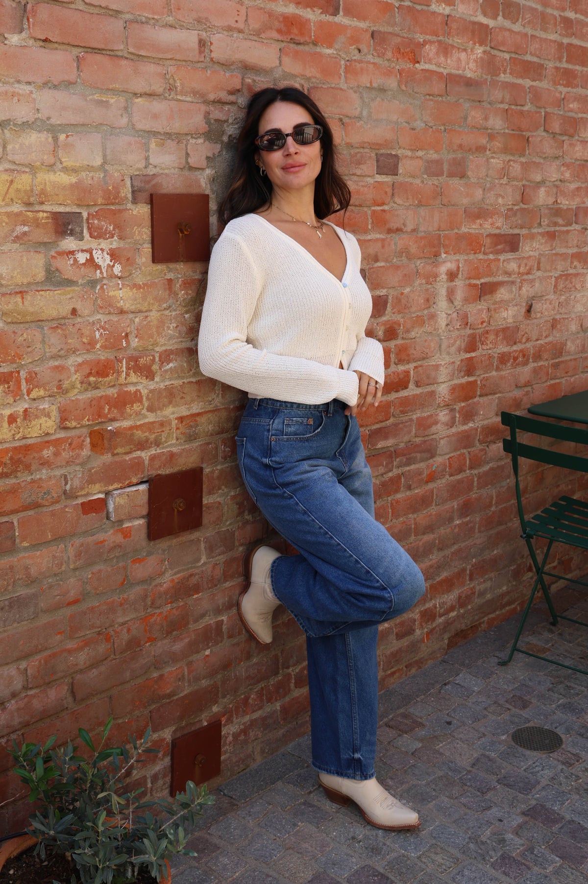Woman in a white blouse and blue jeans leaning against a brick wall.
