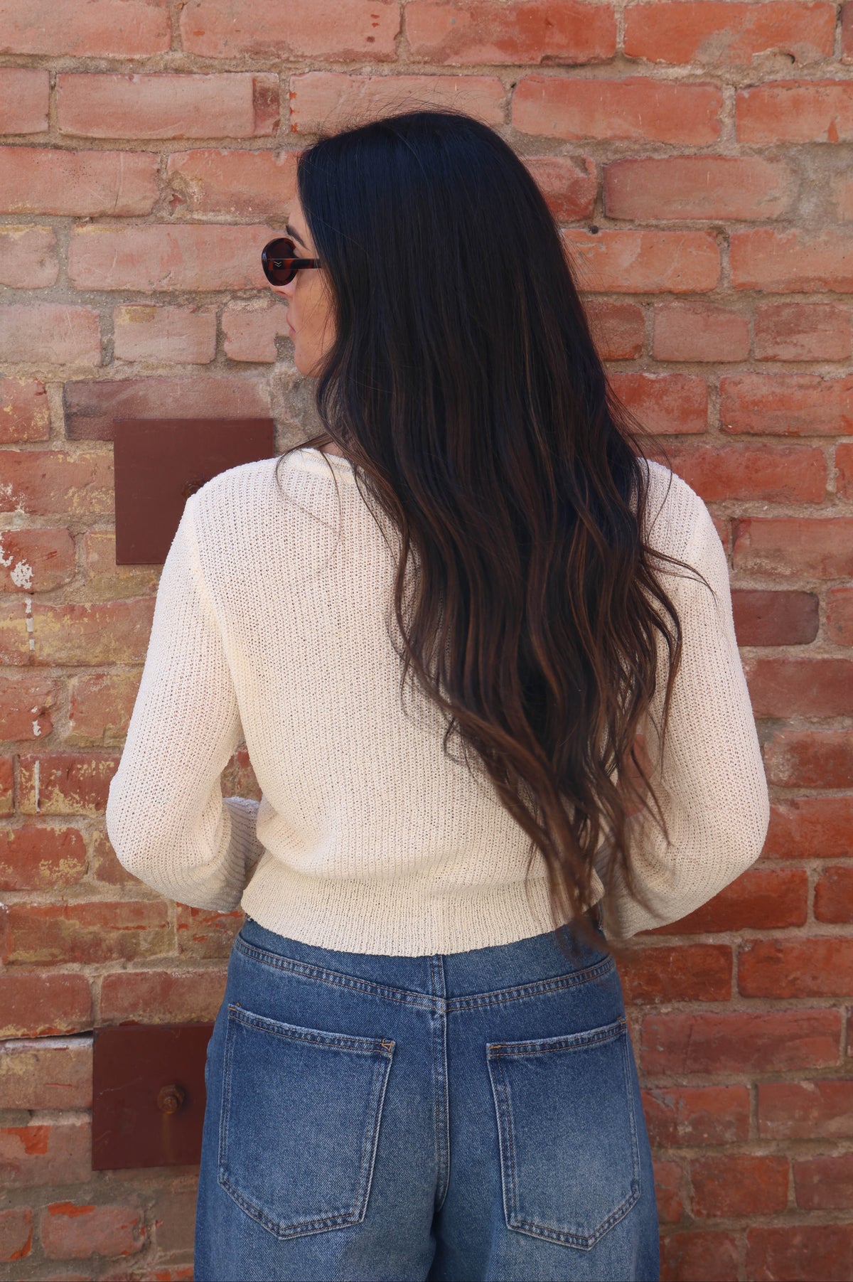 Person wearing a cream sweater and blue jeans standing against a brick wall.