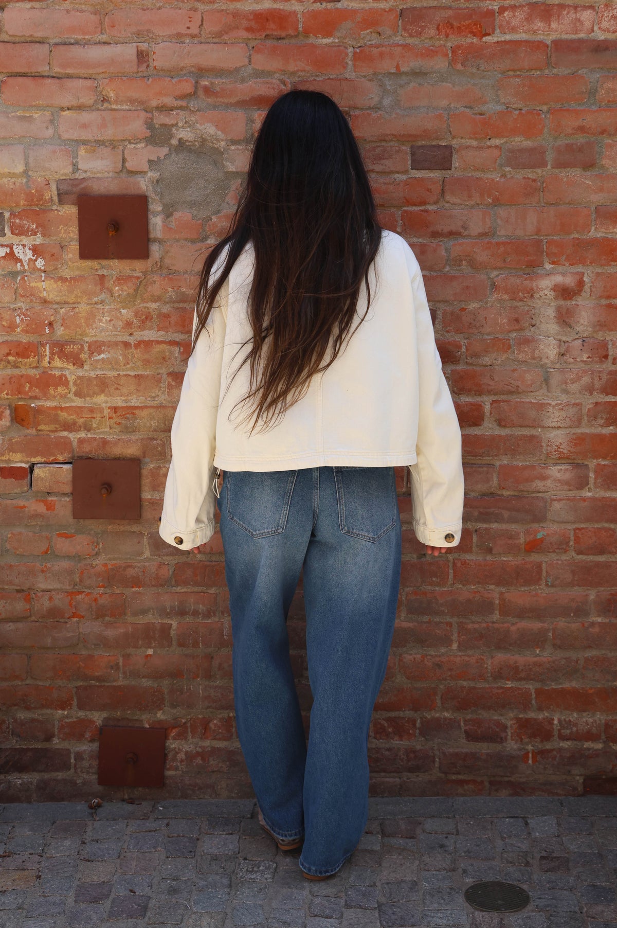 Person wearing a white jacket and blue jeans standing against a brick wall.