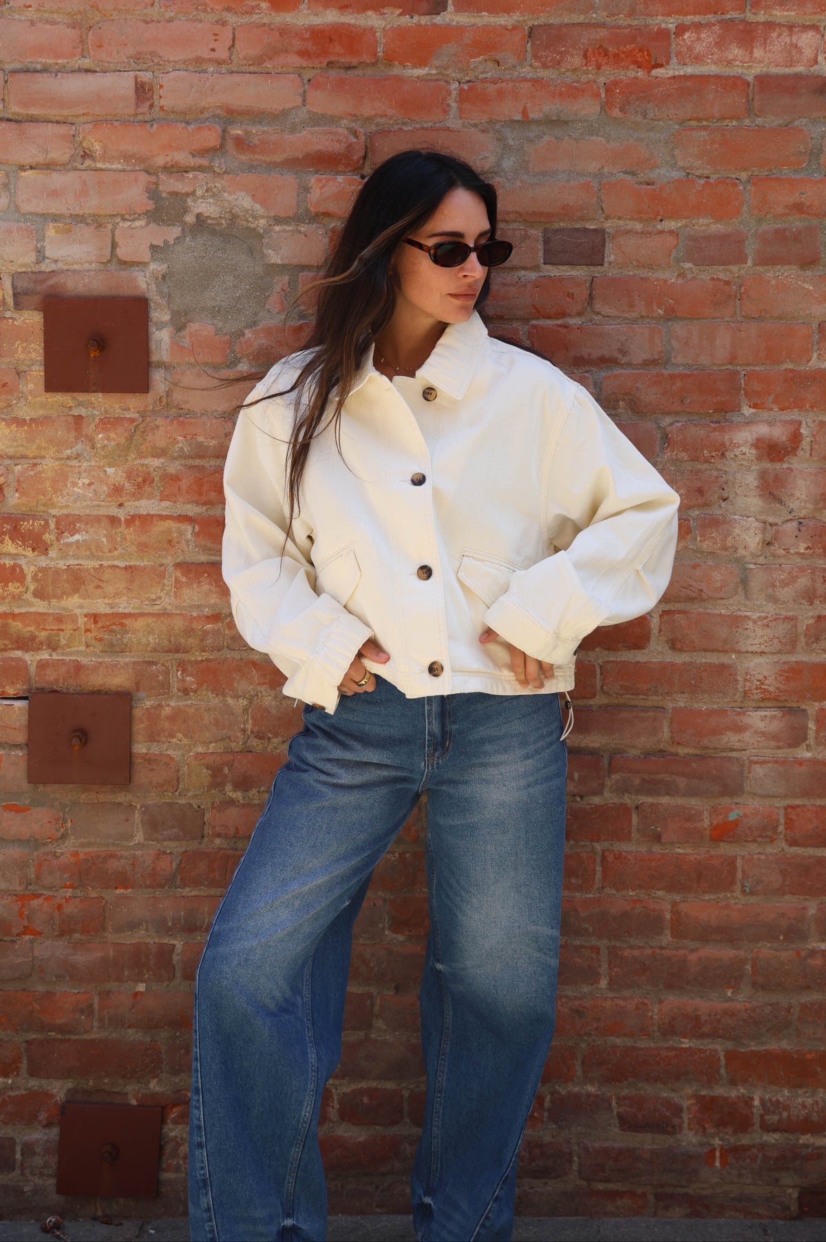 Person wearing a white jacket and blue jeans standing against a brick wall.