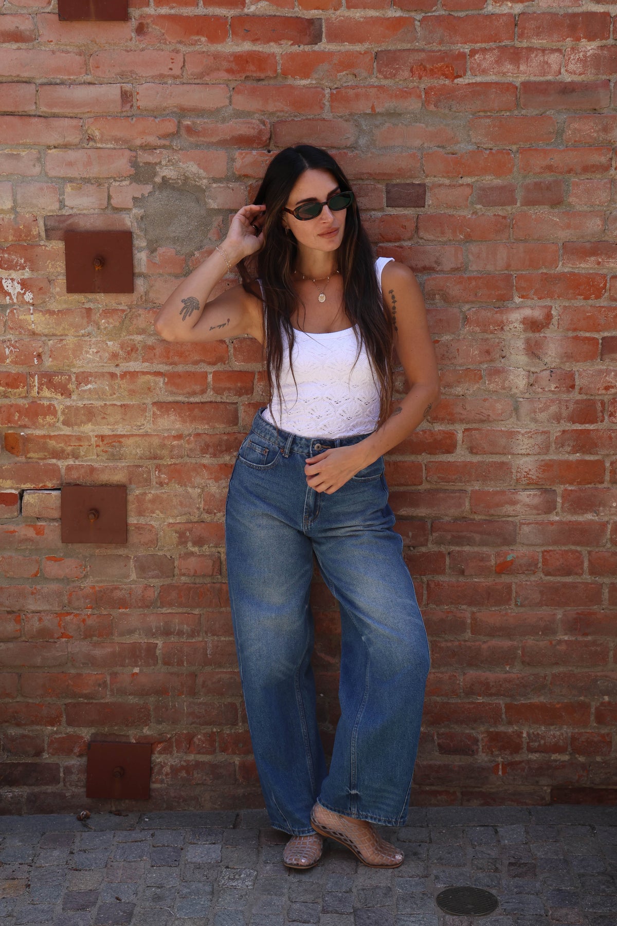 Woman in white top and blue jeans standing against a brick wall.