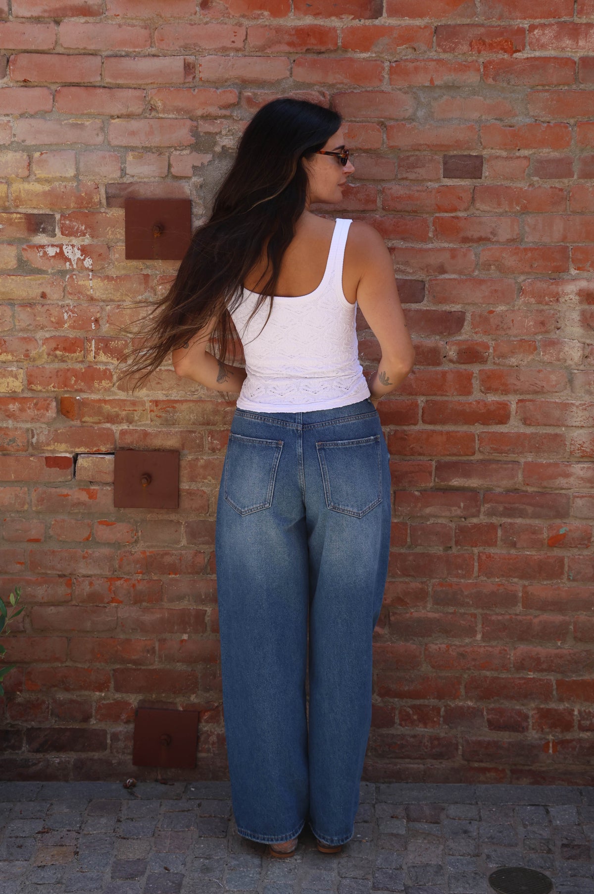 Person wearing a white tank top and blue jeans standing against a brick wall.