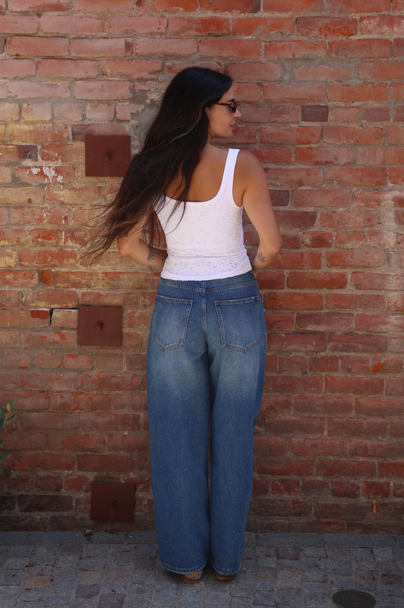 Person wearing a white tank top and blue jeans standing against a brick wall.