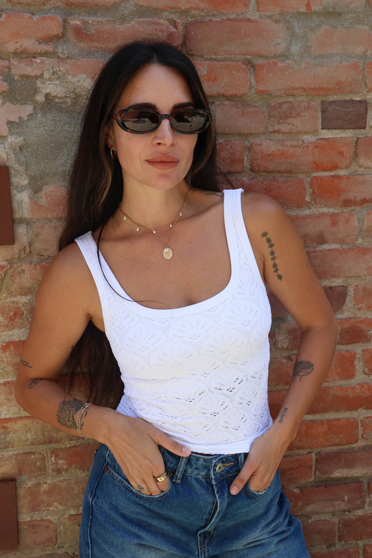 Woman wearing a white tank top and blue jeans against a brick wall.