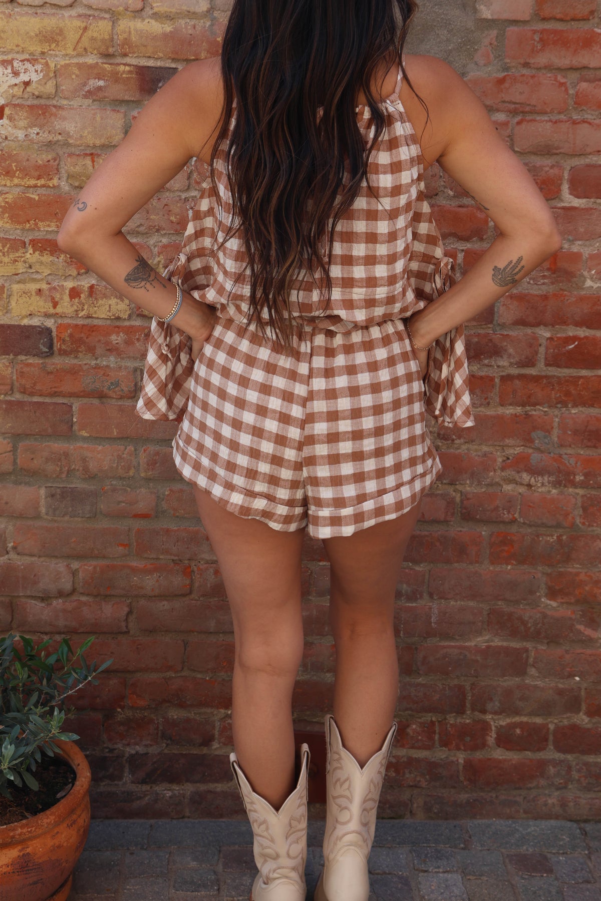 Person wearing a checkered romper and cowboy boots against a brick wall.