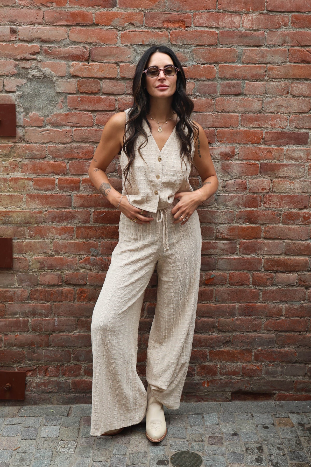 Woman wearing a beige jumpsuit standing against a brick wall.