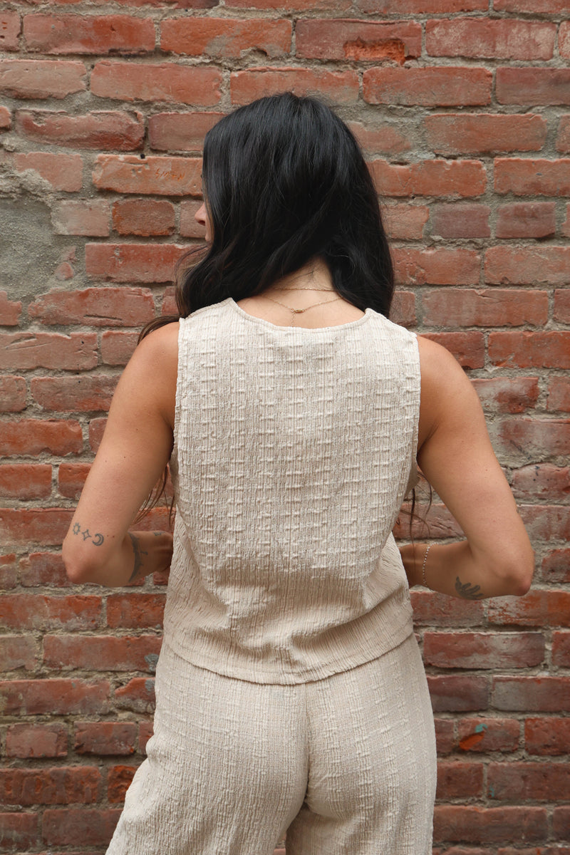 Person wearing a beige sleeveless top and pants against a brick wall.