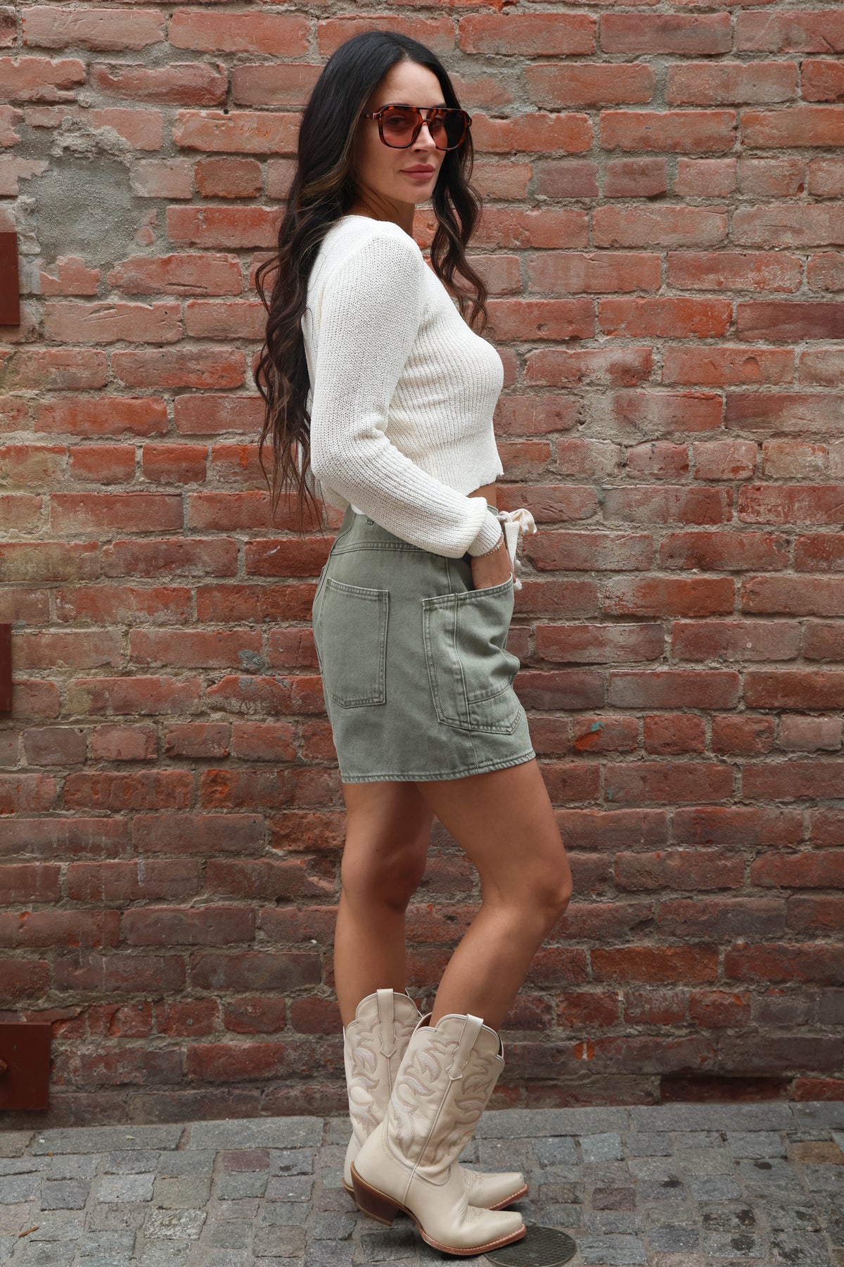Woman wearing a white sweater, green skirt, and white cowboy boots against a brick wall.
