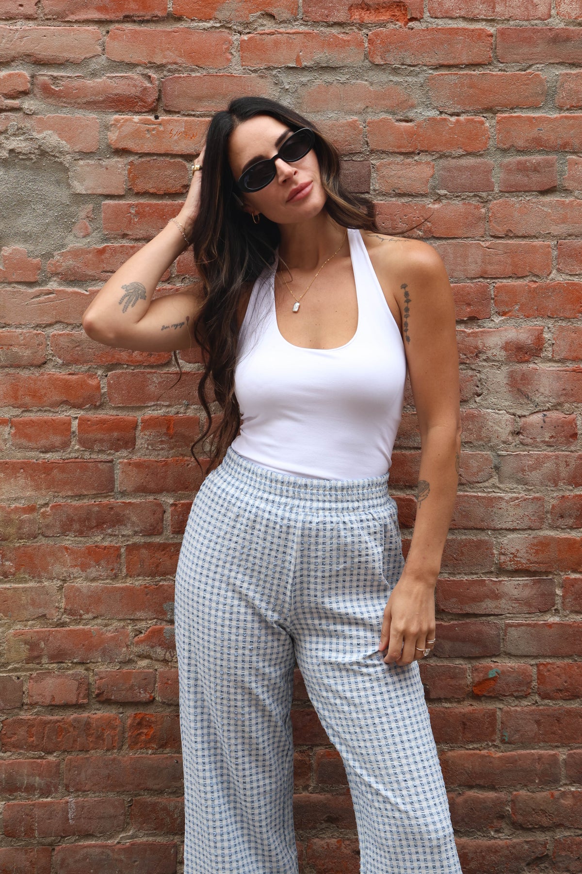 Woman wearing a white tank top and checkered pants against a brick wall.