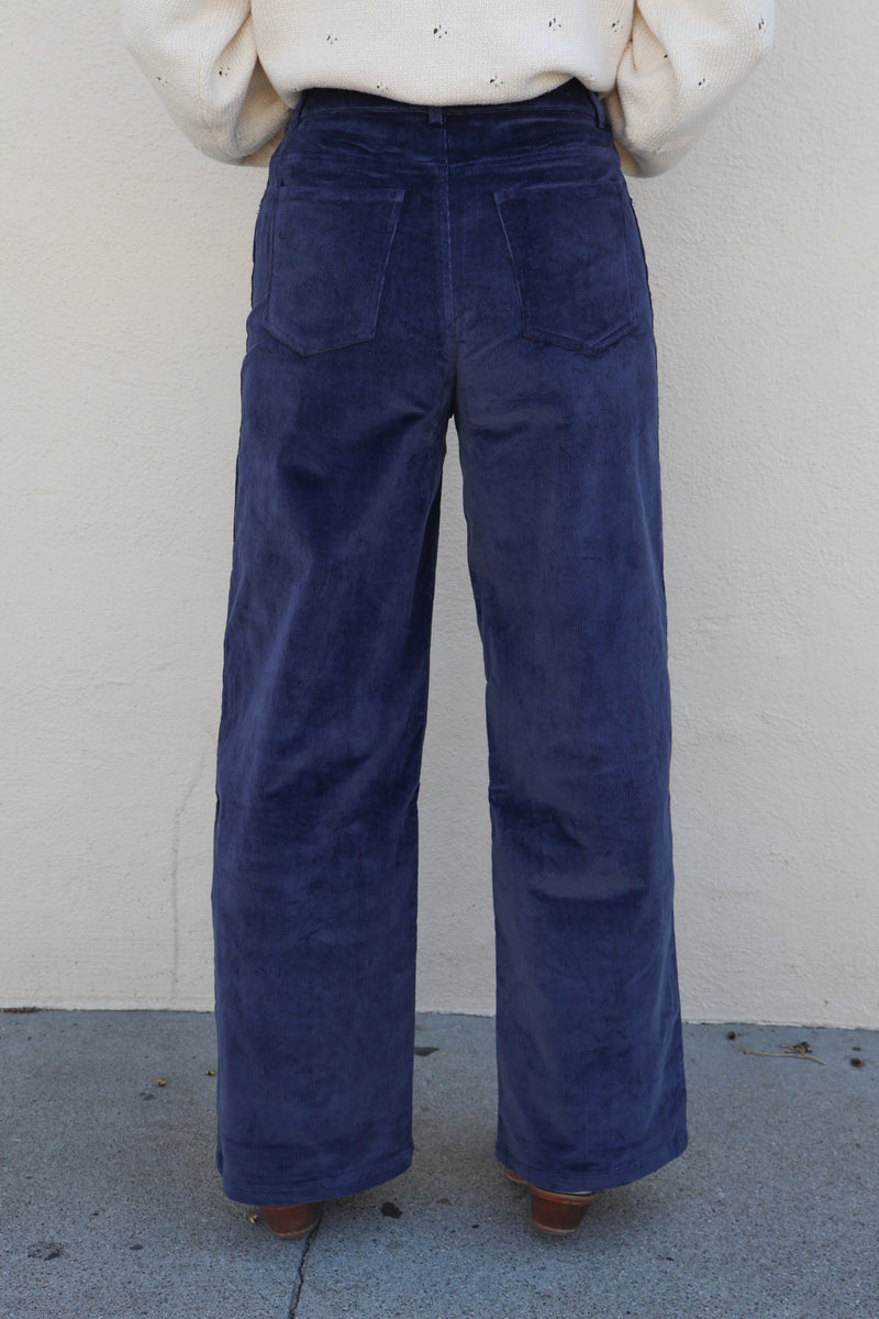 Woman modeling a pair of blue corduroy pants against a plain wall.