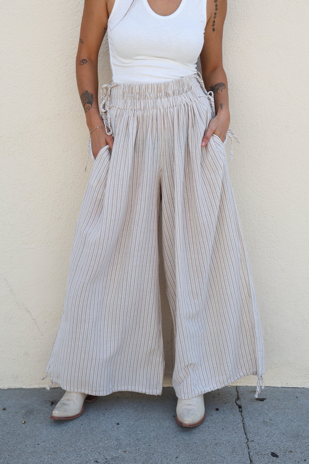 Woman wearing striped flowy pants and a white tank against a plain wall.