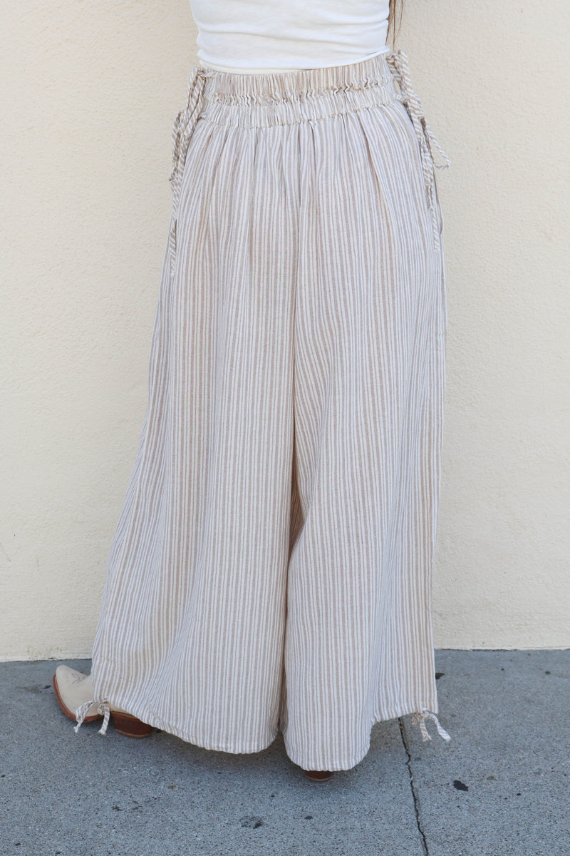 Woman wearing striped flowy pants and a white tank against a plain wall.