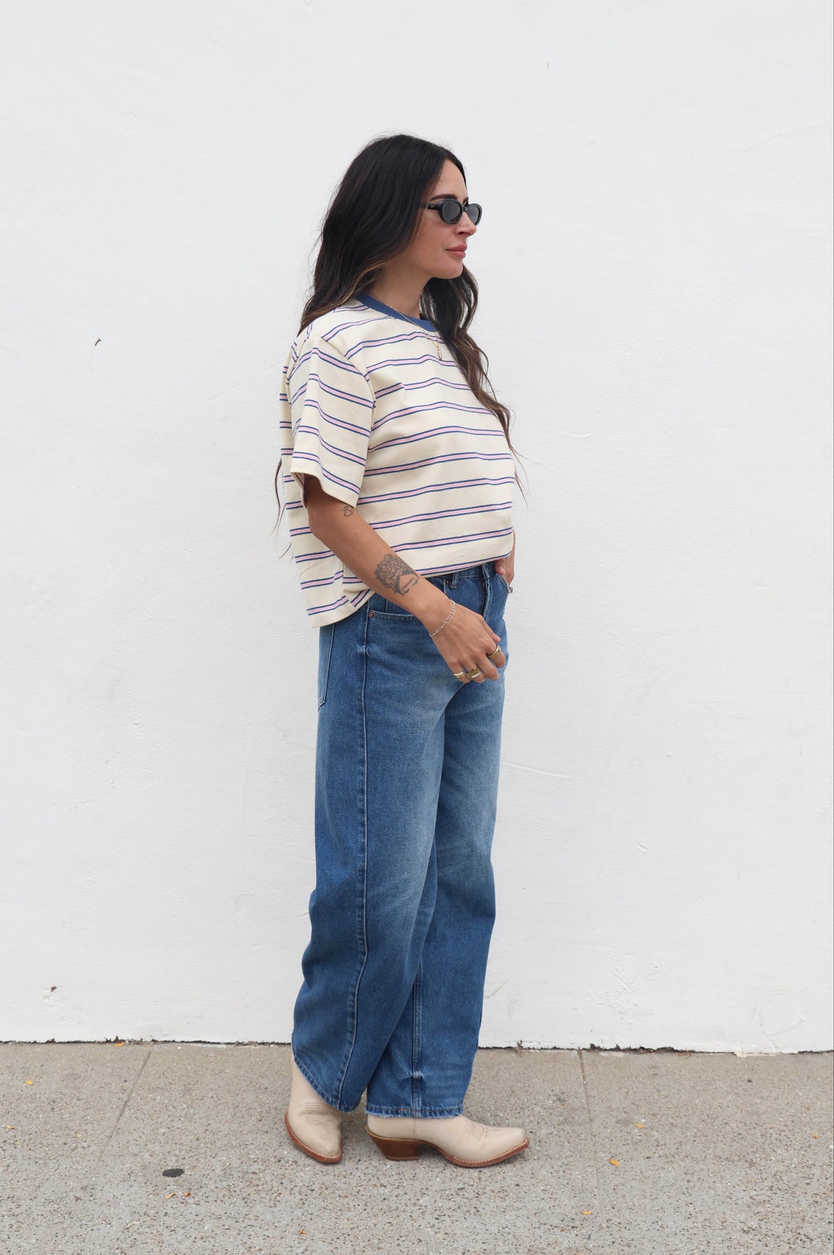 Woman wearing a striped shirt and blue jeans standing against a white wall.