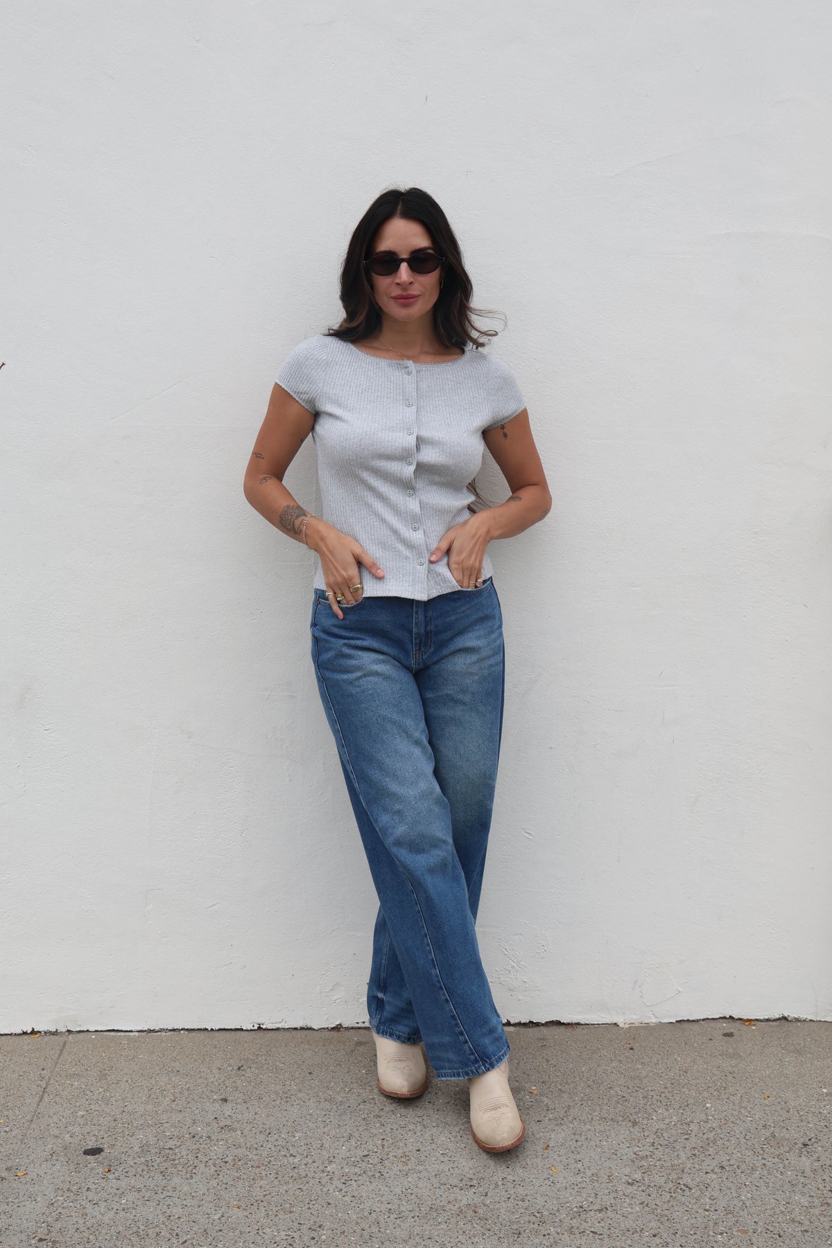Woman wearing a light blue top and jeans standing against a white wall.