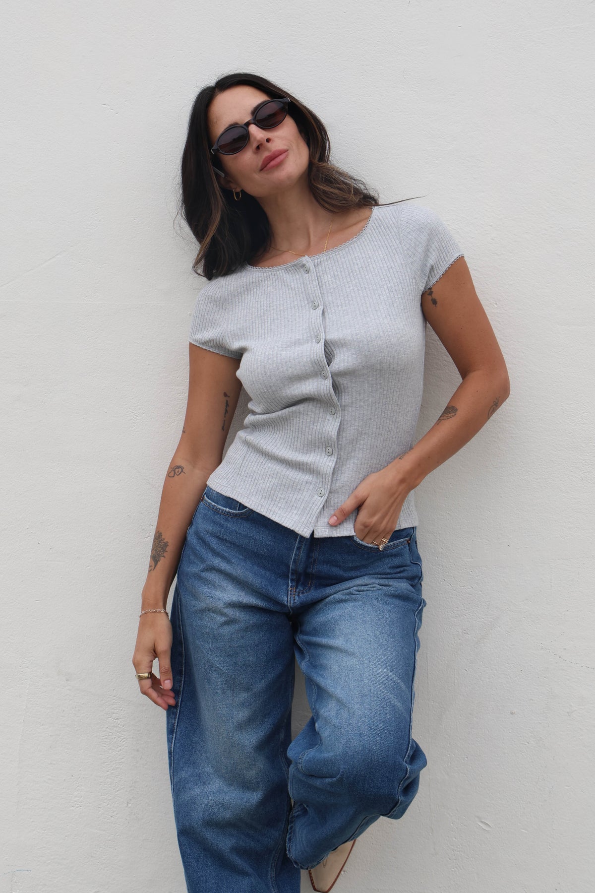 Woman wearing a light gray top and blue jeans against a white wall