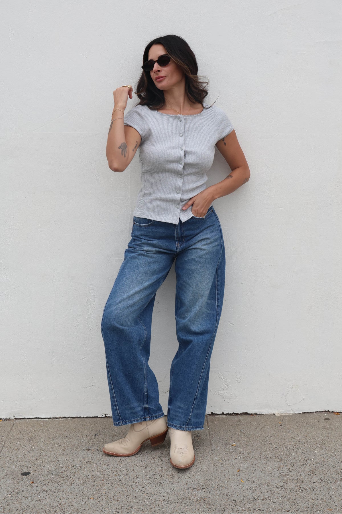 Woman wearing a light gray shirt and blue jeans against a white wall.