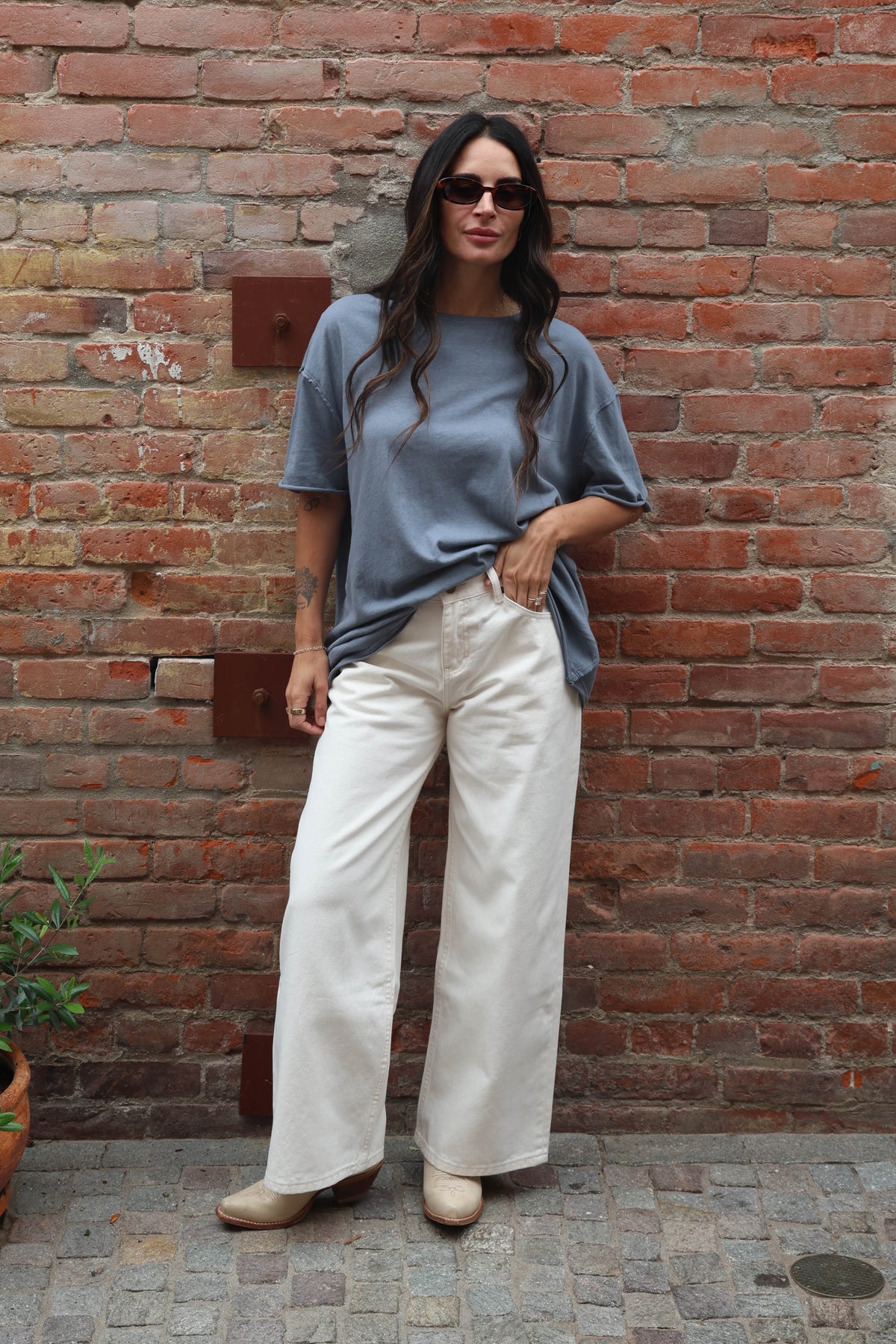 Woman wearing a blue shirt and white pants standing against a brick wall.
