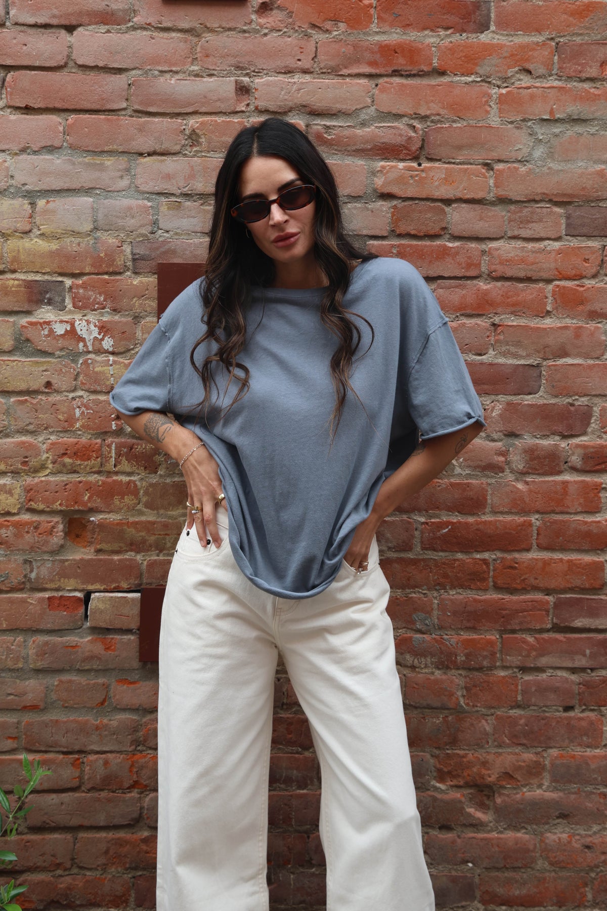 Woman wearing a blue shirt and white pants standing against a brick wall.