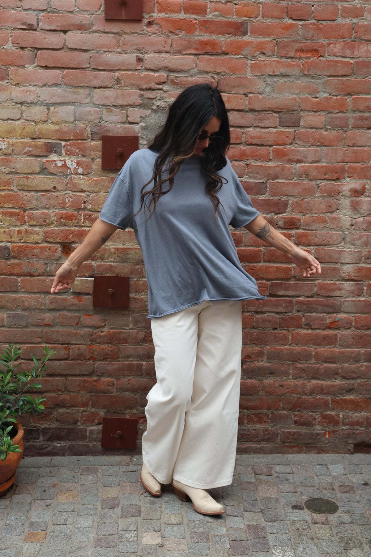 Woman wearing a blue shirt and white pants standing against a brick wall.