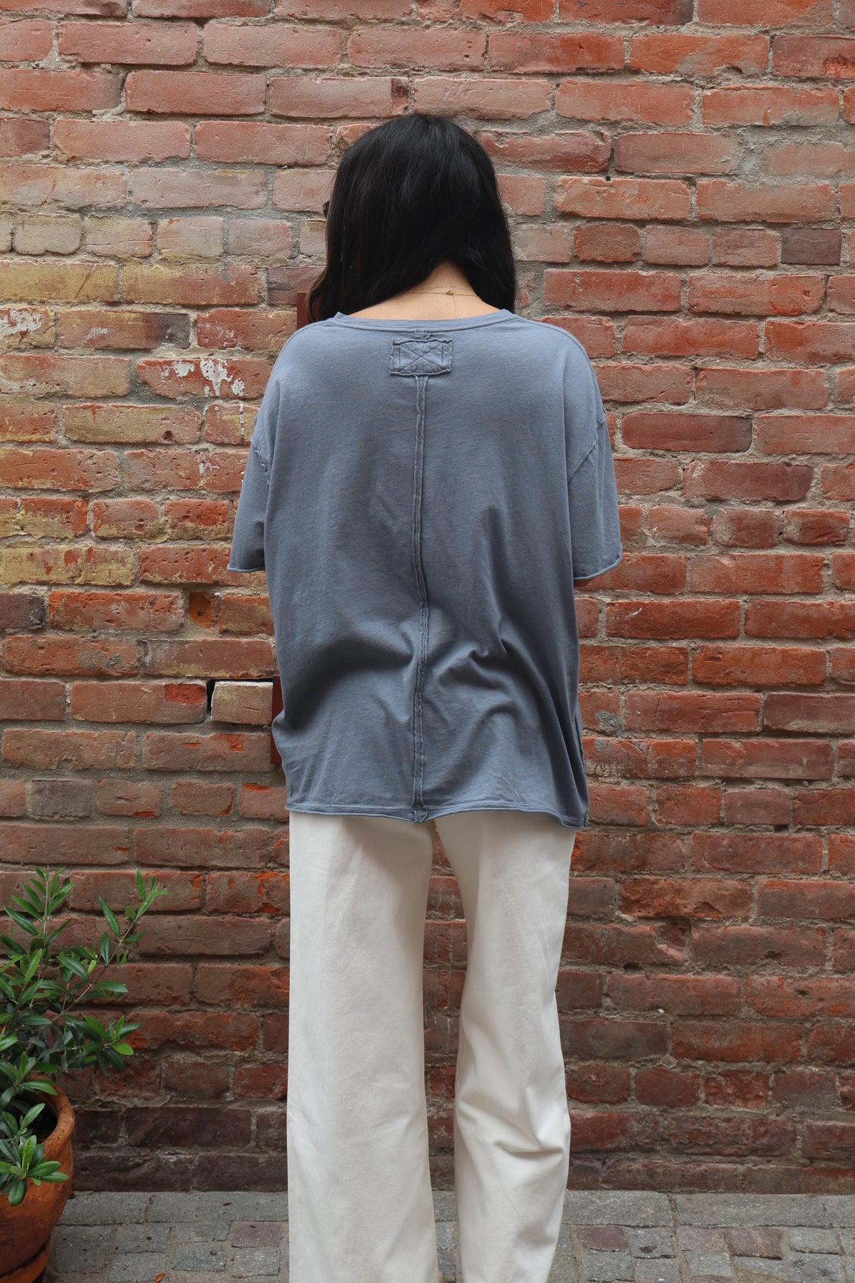 Person wearing a gray oversized shirt and white pants standing against a brick wall.
