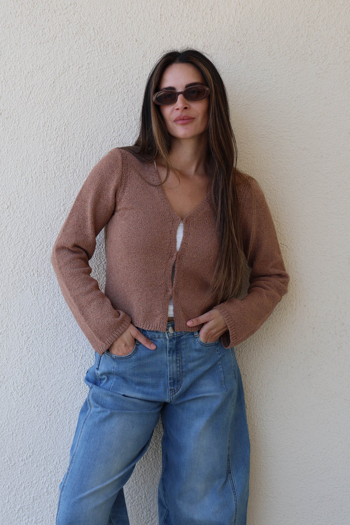 Woman wearing a toffee knit cardigan with jeans against a plain wall.