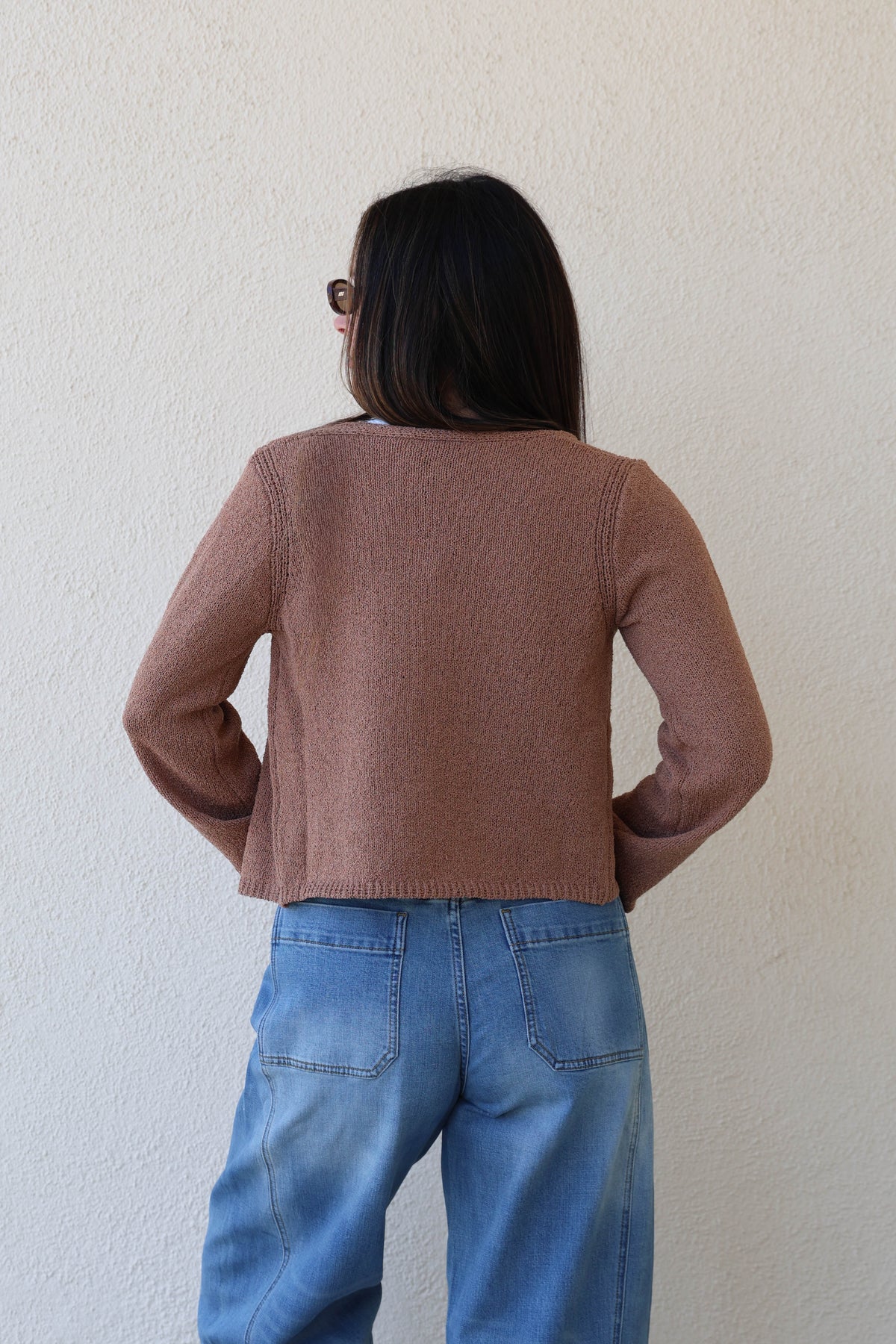 Woman wearing a toffee knit cardigan with jeans against a plain wall.