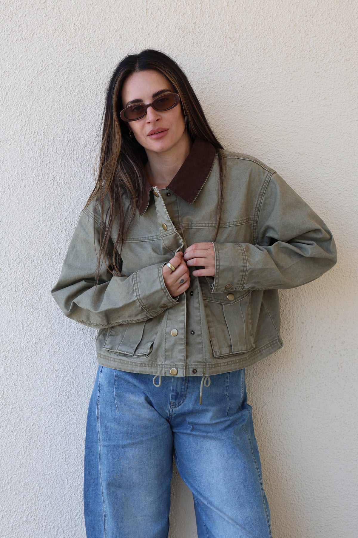 Woman wearing a green utility jacket and jeans against a white wall.
