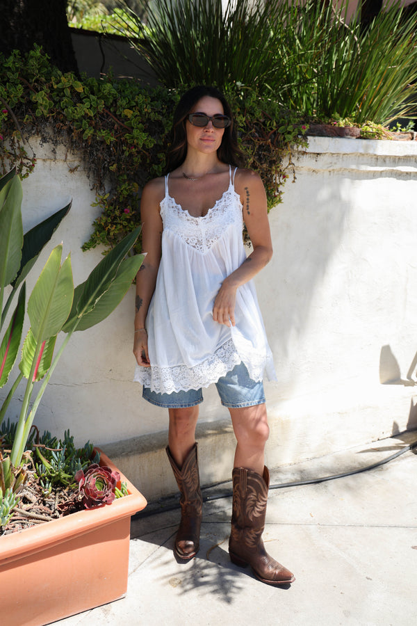 Woman in a white dress and cowboy boots standing outdoors with plants and a wall in the background