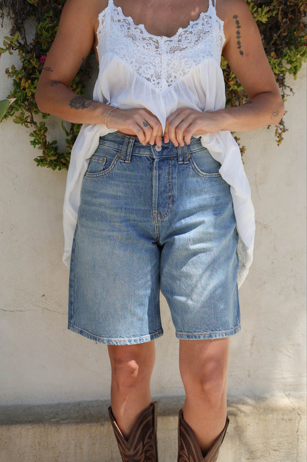 Person wearing a white lace top, denim shorts, and brown cowboy boots against a neutral wall.