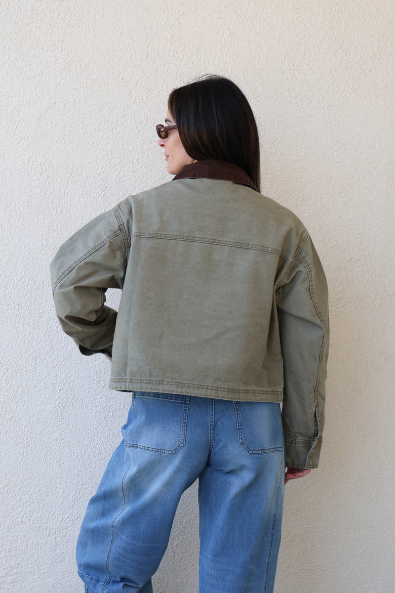 Woman wearing a green utility jacket and jeans against a white wall.
