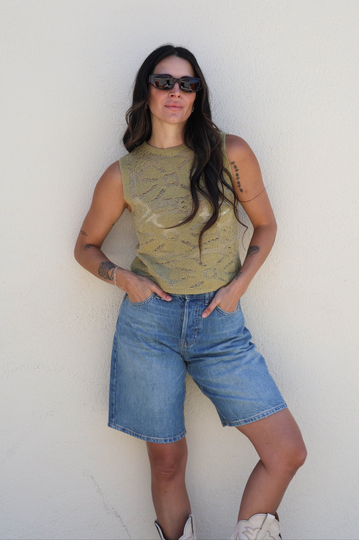 Woman wearing a green sleeveless top and blue shorts against a white wall.