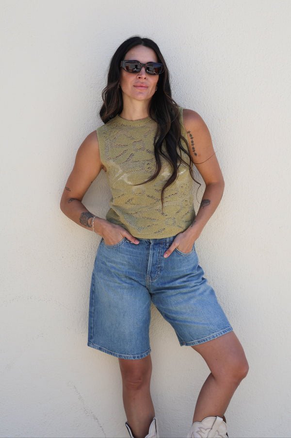 Woman wearing a green sleeveless top and blue shorts against a white wall.