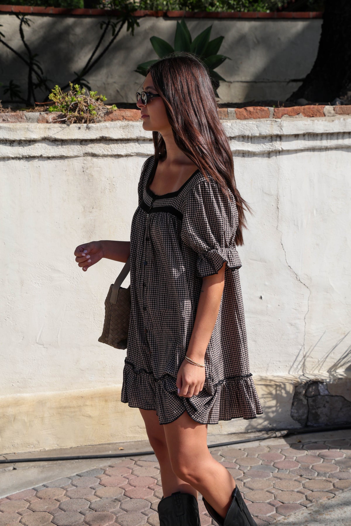 Woman wearing a brown and black gingham dress with black boots against a white wall.