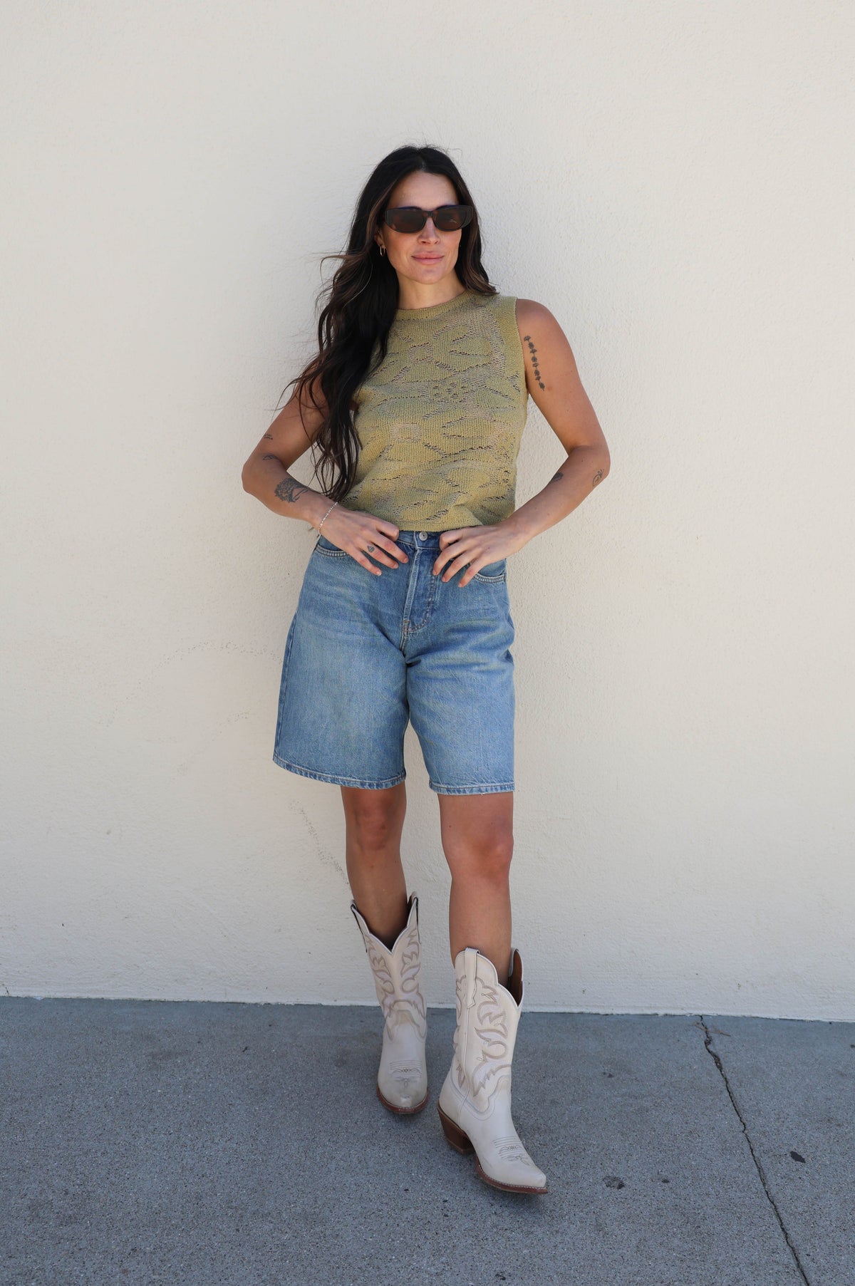 Woman wearing a sleeveless top, denim shorts, and cowboy boots against a white wall.