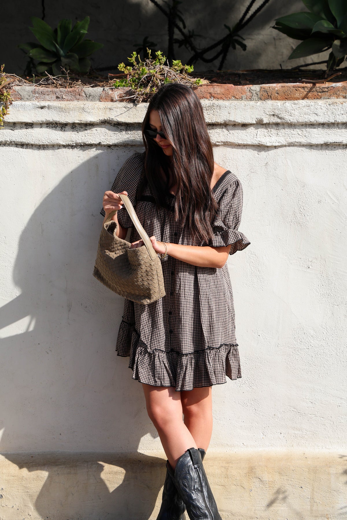 Woman wearing a brown and black gingham dress with black boots against a white wall.
