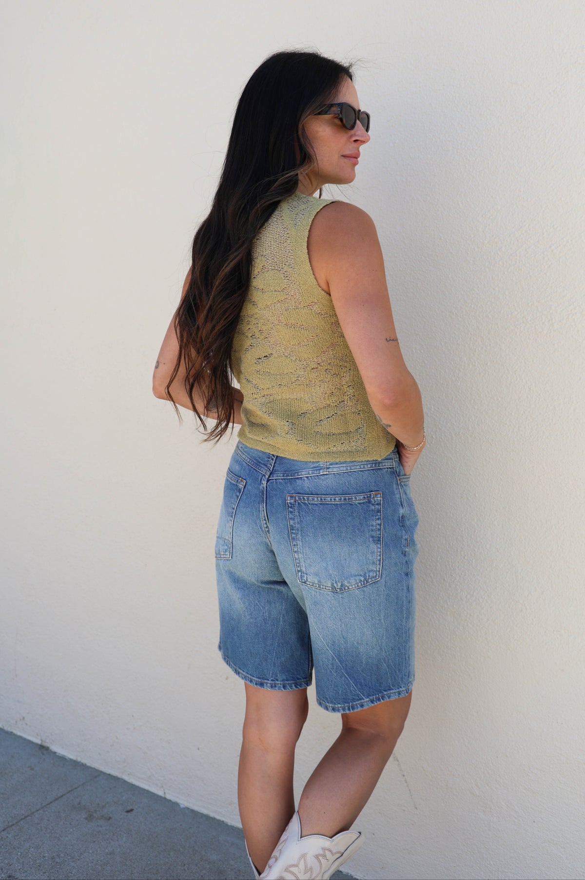 Woman wearing a green sleeveless top and blue denim shorts against a white wall.