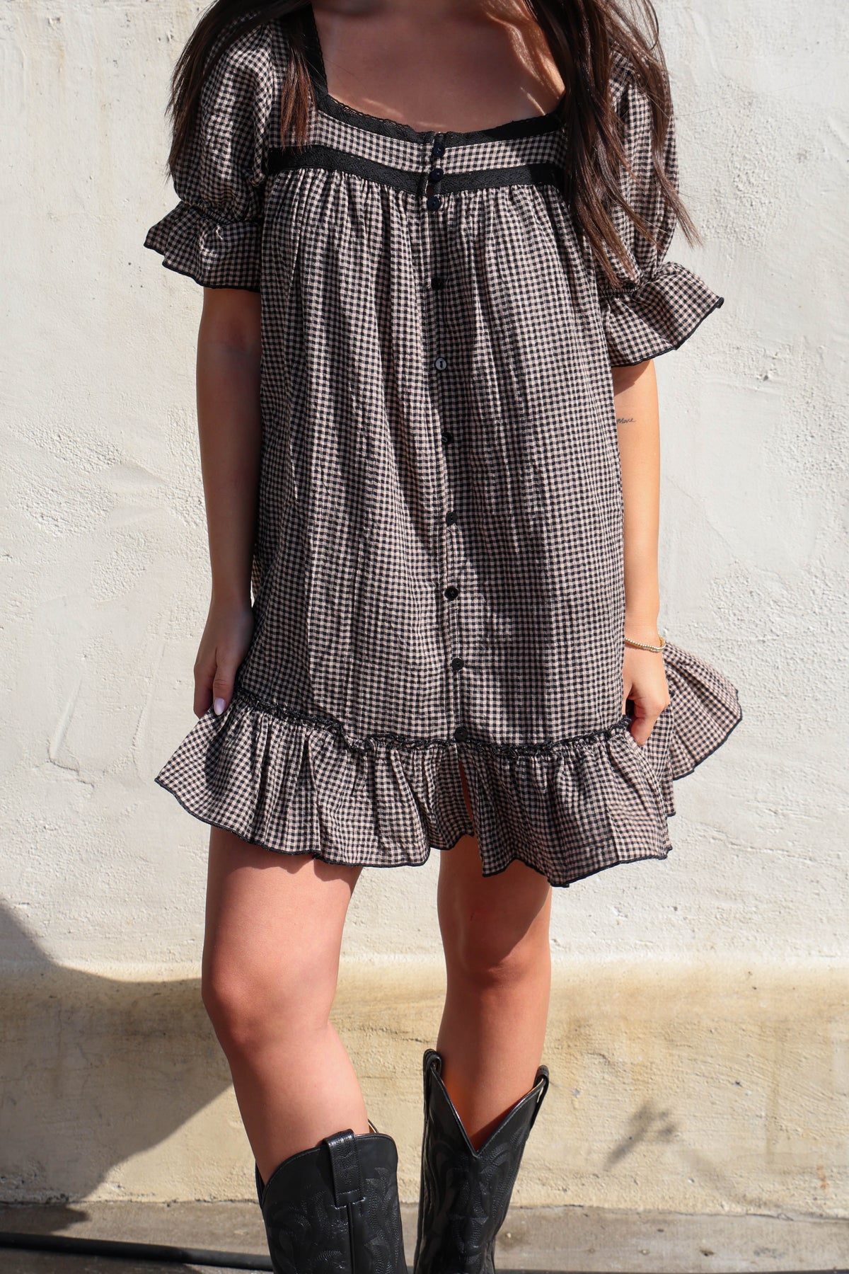 Woman wearing a brown and black gingham dress with black boots against a white wall.