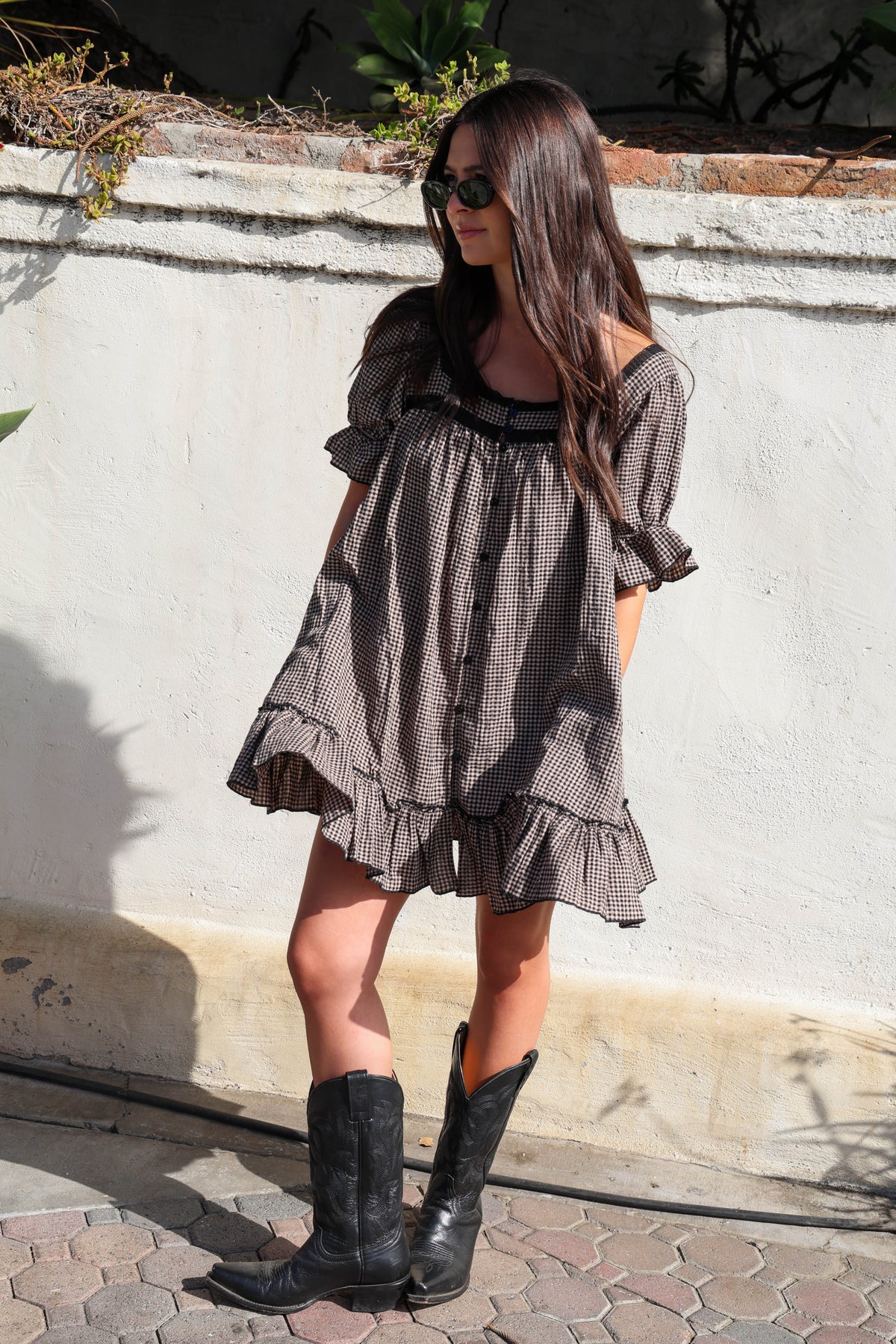 Woman wearing a brown and black gingham dress with black boots against a white wall.