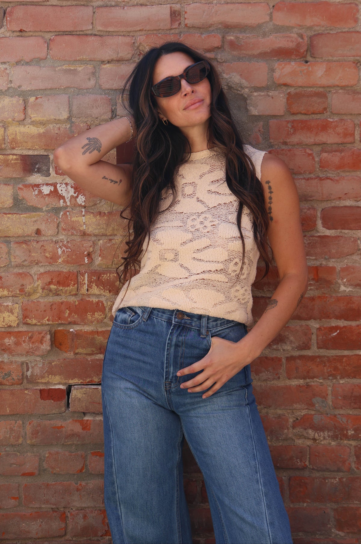 Woman wearing sunglasses and a beige top against a brick wall
