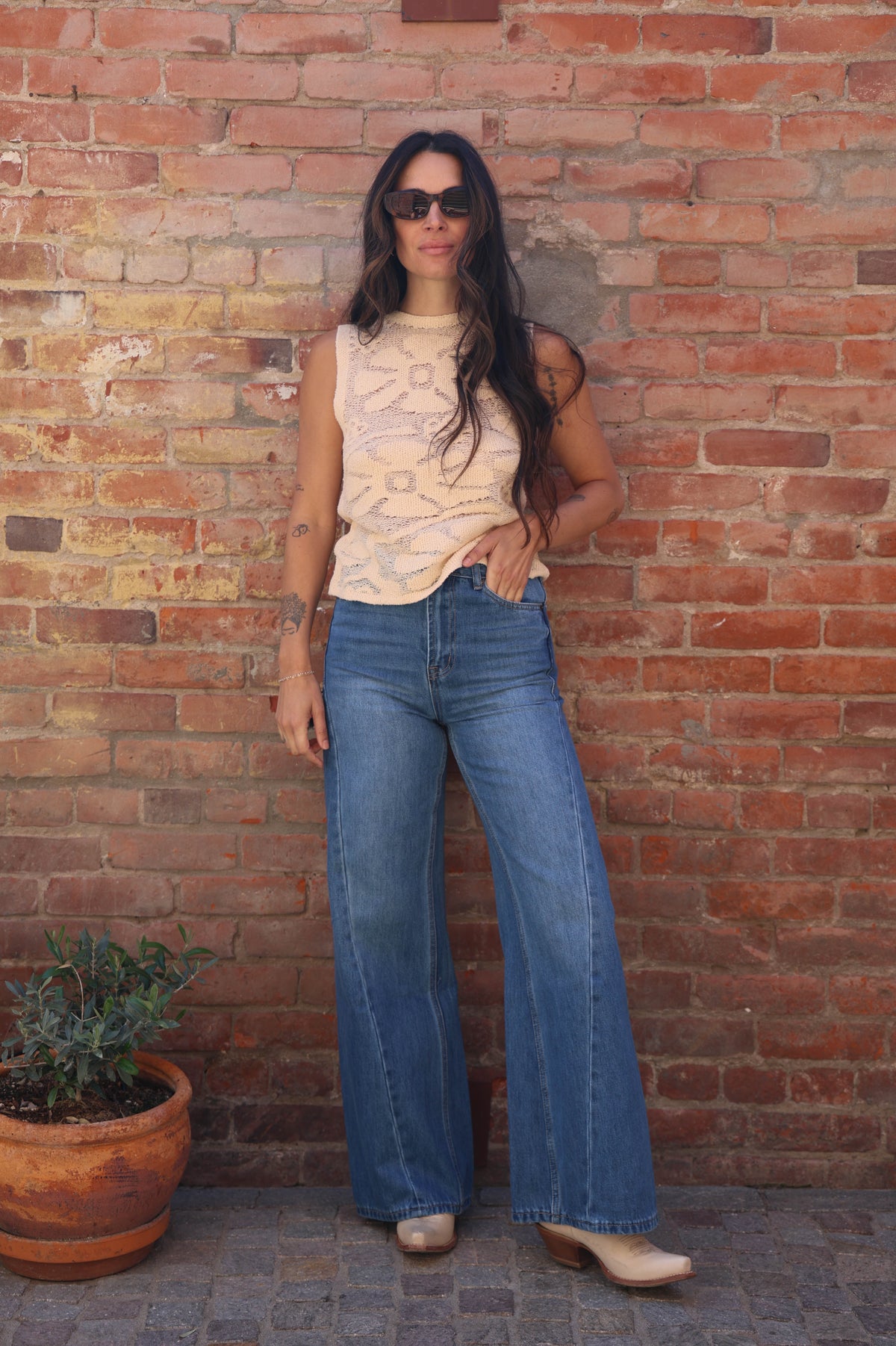 Woman in a sleeveless top and blue jeans standing against a brick wall.