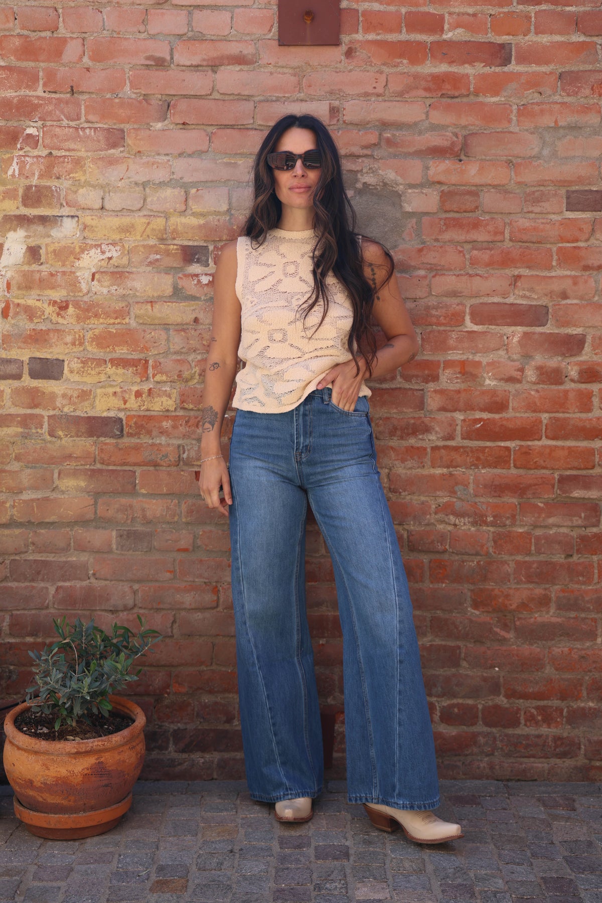 Woman in a sleeveless top and blue jeans standing against a brick wall.