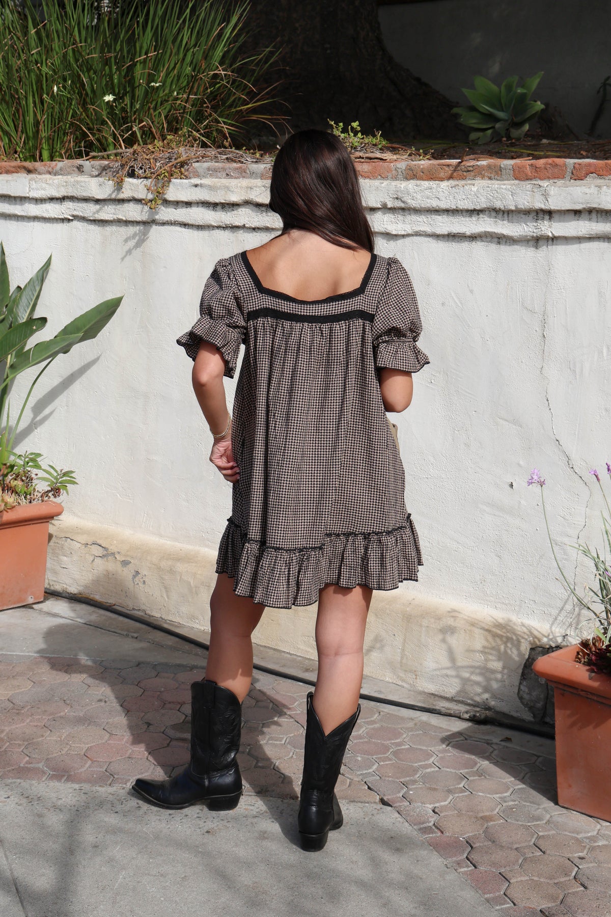 Woman wearing a brown and black gingham dress with black boots against a white wall.