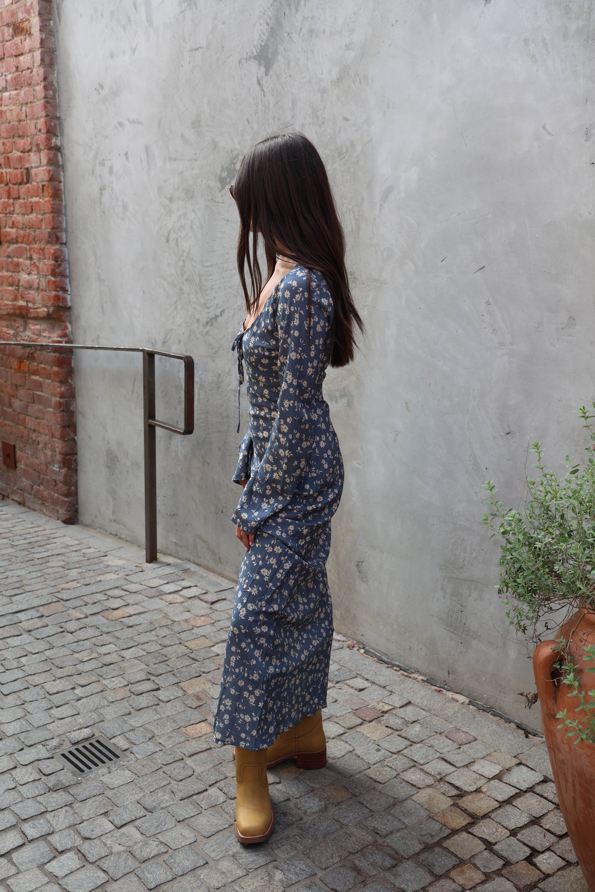 A woman wearing a long-sleeve blue floral maxi dress and brown boots stands against a textured gray concrete wall next to a potted plant.