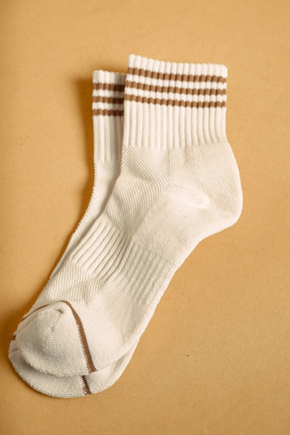 A pair of off-white crew socks with brown stripes near the cuff, laid flat on a warm tan background.
