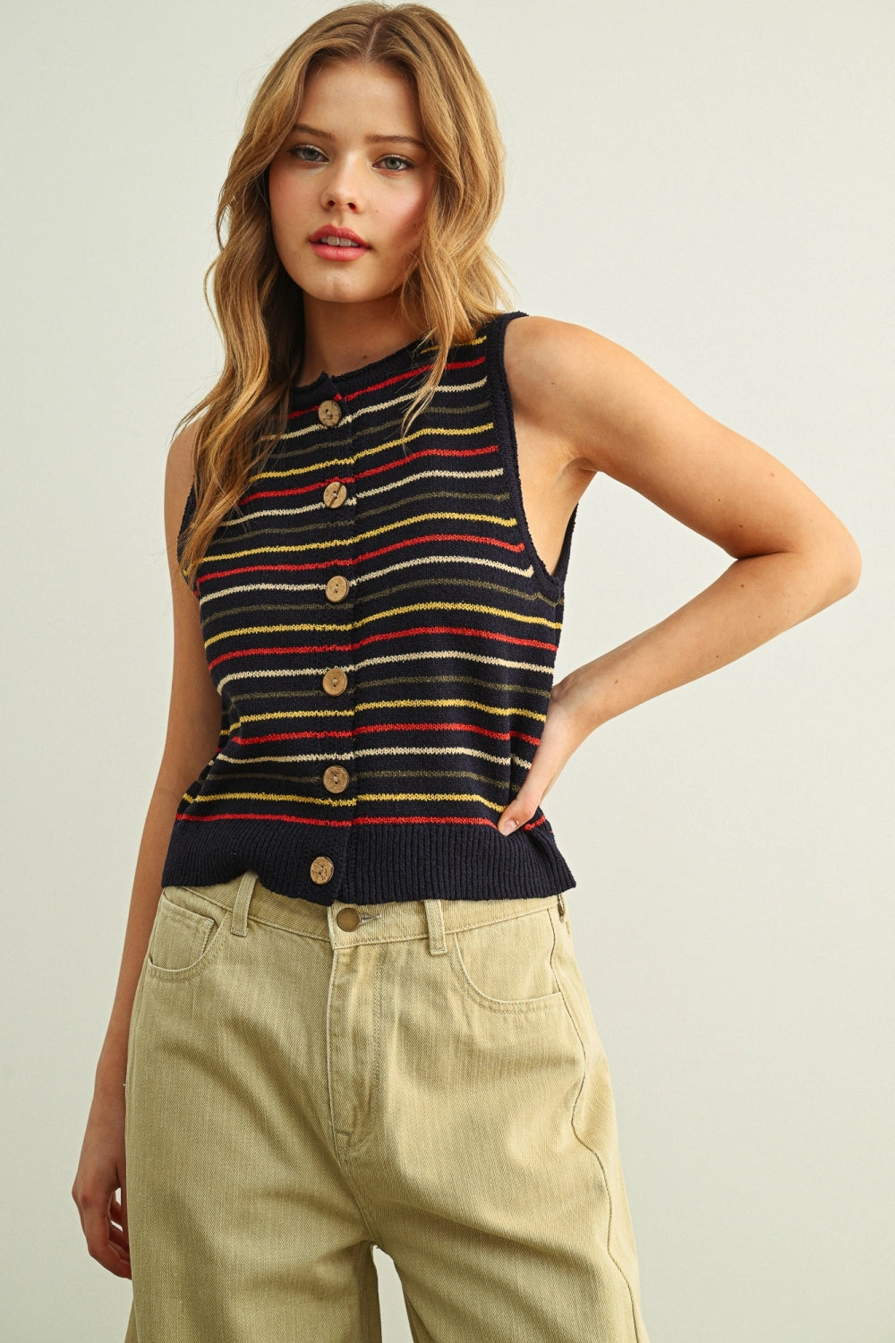 Woman wearing a striped sleeveless top with buttons and beige pants on a plain background