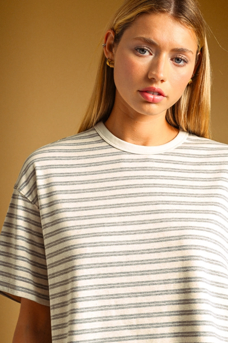 A close-up shot of a woman wearing a short-sleeve crewneck t-shirt with thin horizontal gray stripes on a cream-colored fabric, set against a solid warm tan background.